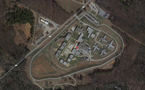 Aerial view of a large facility surrounded by trees and roads, with multiple buildings and open areas visible.