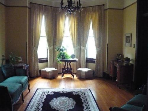 A cozy room with large windows, elegant curtains, a round table, and vintage furniture on a patterned rug.