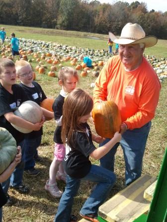 A man in a cowboy hat helps a girl lift a pumpkin while children watch in a pumpkin patch.