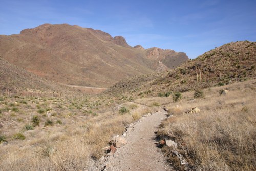 A winding dirt path leads through a dry, mountainous landscape with sparse vegetation under a clear blue sky.