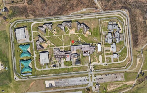 Aerial view of a large facility with multiple buildings and a surrounding fence, set in a green landscape.