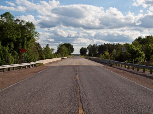 A wide, empty road stretches into the distance, flanked by greenery and under a partly cloudy sky.