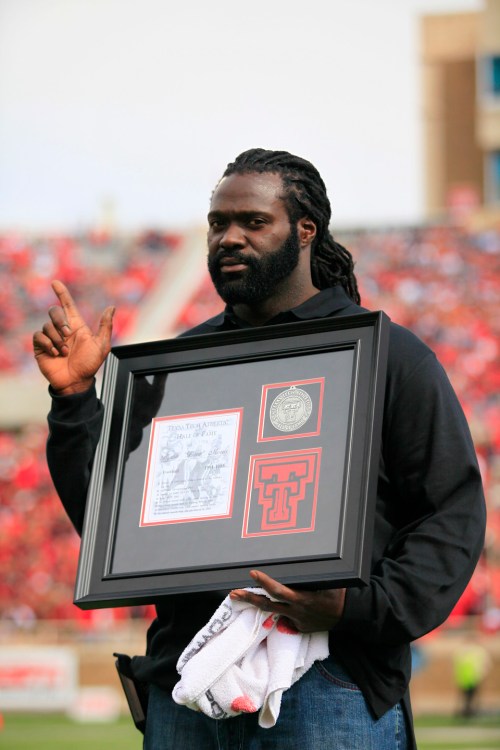 A man holds a framed award while making a peace sign, standing on a football field with a crowd in the background.