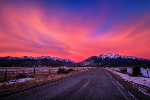 A scenic road leads towards majestic mountains under a vibrant sunset sky with pink and purple hues.