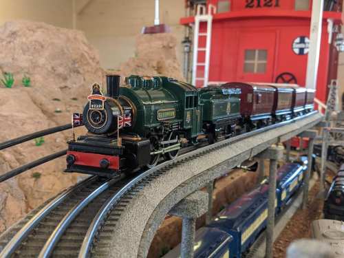 A model steam train on curved tracks, passing by a rocky landscape and a red train station in the background.