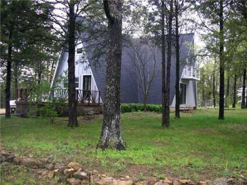 A modern A-frame house nestled among tall trees, with a wooden deck and green grass surrounding it.