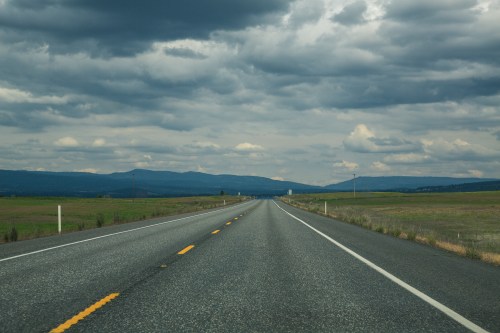 A long, empty road stretches into the distance under a cloudy sky, with mountains visible on the horizon.