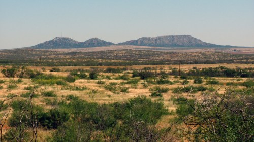 A panoramic view of three distant mountains against a clear sky, surrounded by open grassland and sparse vegetation.