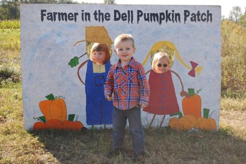 A young child stands in front of a painted backdrop featuring cartoon farmers and pumpkins in a pumpkin patch.