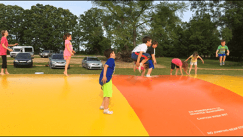 Children playing on a large inflatable bounce pad outdoors, with trees and cars in the background.