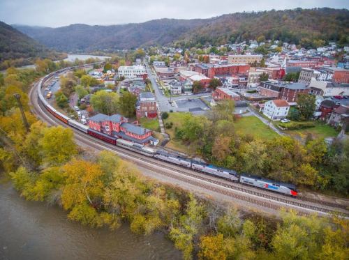 Aerial view of a train traveling along a river, with a small town and autumn foliage in the background.