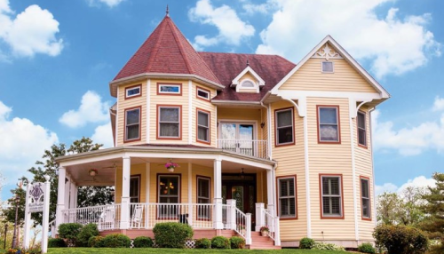 A charming two-story yellow house with a turret, white railings, and a porch, set against a blue sky with clouds.