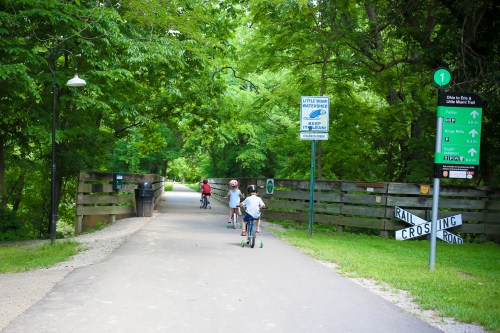 Children riding bikes on a tree-lined path, with signs for a bike trail and a railroad crossing ahead.