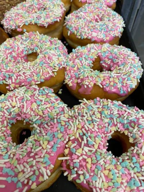 A close-up of freshly baked donuts topped with pink icing and colorful sprinkles, arranged on a tray.