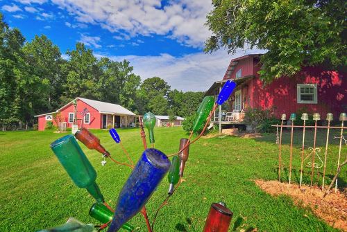 A colorful bottle tree in the foreground with a red barn and green trees under a blue sky in the background.
