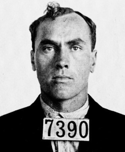 Black and white mugshot of a man with short hair, wearing a suit, displaying the number 7390.