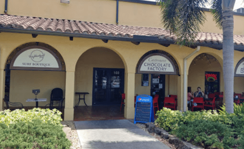 Exterior of a building featuring a surf boutique and a chocolate factory, with outdoor seating and tropical landscaping.