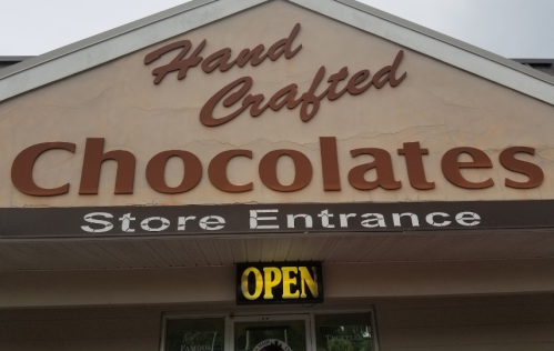 Sign for a chocolate store featuring "Hand Crafted Chocolates" and an illuminated "OPEN" sign.