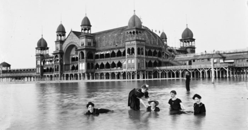 Women in vintage swimwear wade in shallow water near a grand seaside building with ornate architecture.