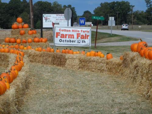 A farm fair sign surrounded by pumpkins and hay bales, with a road and trees in the background.
