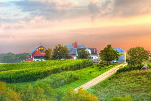 A picturesque farm at sunset, featuring red barns, green fields, and a winding dirt road.