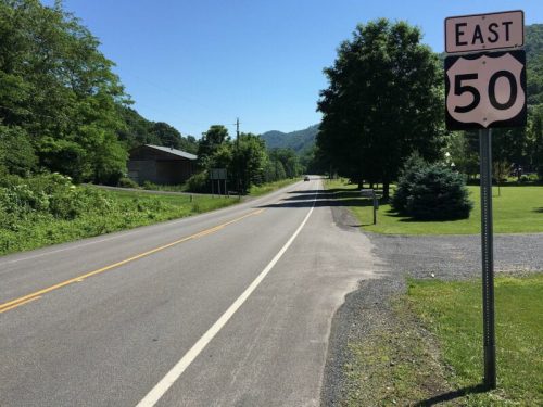 A clear road sign for Route 50 East beside a quiet rural road lined with trees and mountains in the background.