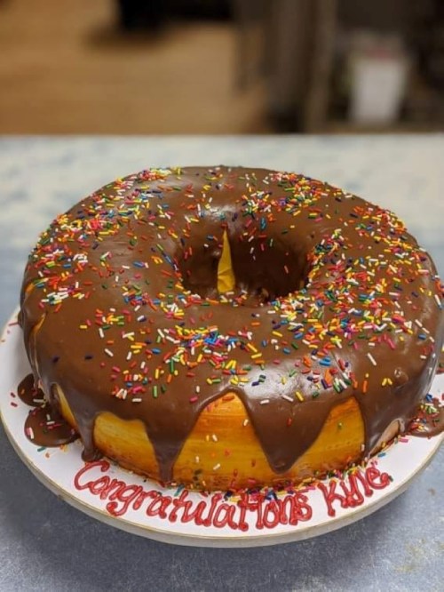 A large, round chocolate-covered donut cake with colorful sprinkles and "Congratulations Kate" written in red icing.