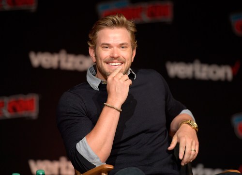 A smiling man in a dark sweater sits on a stage, holding his chin, with a Verizon backdrop behind him.