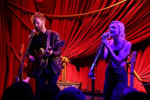A male musician plays guitar while a female singer performs, both on stage with red drapery in the background.