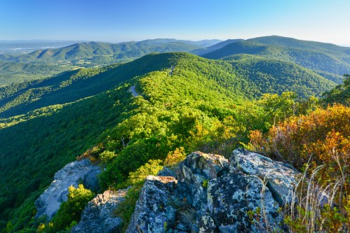 A panoramic view of lush green mountains under a clear blue sky, with rocky outcrops in the foreground.