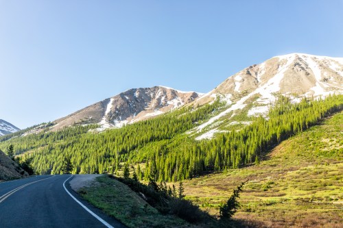 A winding road leads through lush green forests and snow-capped mountains under a clear blue sky.