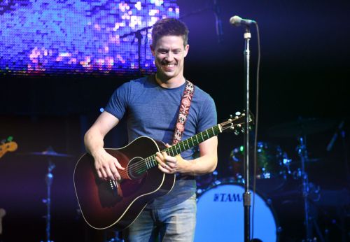 A smiling musician plays an acoustic guitar on stage, with colorful lights and a drum set in the background.
