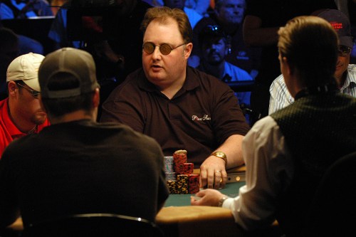 A group of poker players at a table, focused on the game, with one player wearing glasses and a dark shirt.