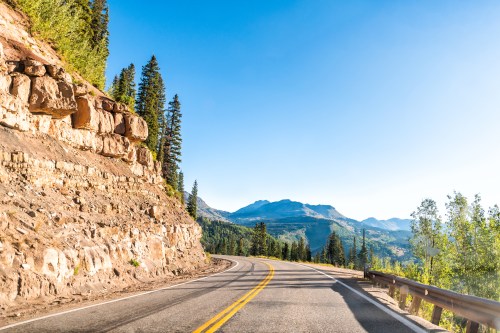 A winding road curves through a mountainous landscape under a clear blue sky, surrounded by trees and rocky terrain.