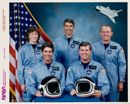 A group portrait of five NASA astronauts in blue jumpsuits, with an American flag and space shuttle in the background.