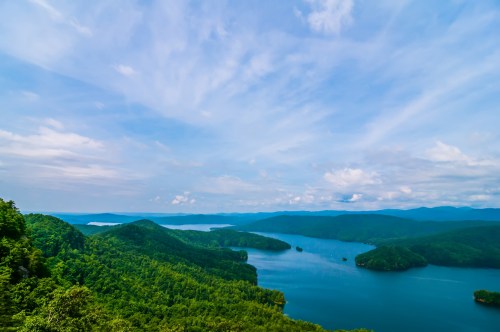 A panoramic view of a lush green landscape with a serene lake and blue sky dotted with clouds.