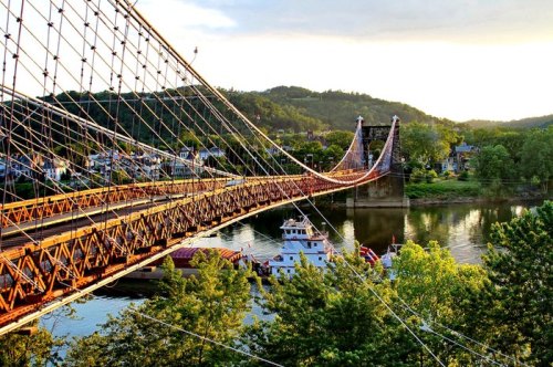 A suspension bridge spans a river, surrounded by lush greenery and a small town in the background under a golden sunset.