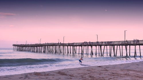 A surfer rides a wave near a wooden pier at sunset, with people walking along the pier under a pastel sky.
