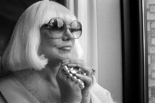 A glamorous person with white hair and oversized sunglasses gazes thoughtfully out a window, holding a pearl bracelet.