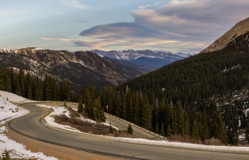 A winding road through snow-covered mountains and evergreen trees under a colorful sky.