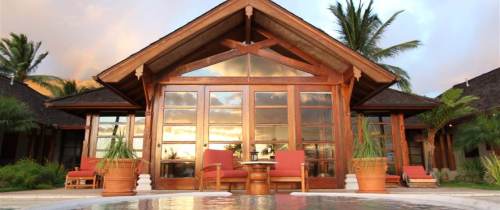 A wooden house with large glass windows, surrounded by palm trees and a pool, set against a colorful sunset.