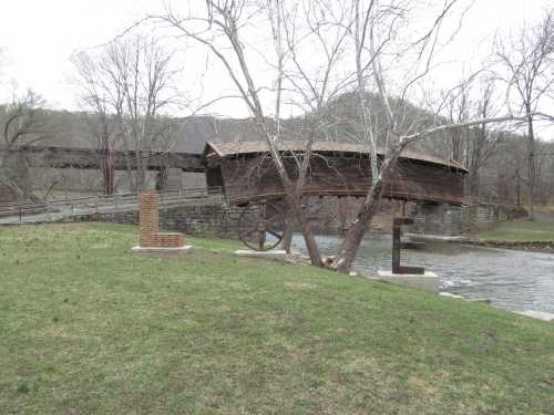 A wooden covered bridge spans a river, surrounded by bare trees and grassy banks.