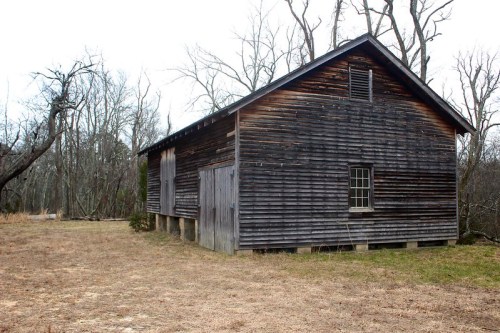 A weathered wooden barn stands in a sparse, grassy area surrounded by bare trees.