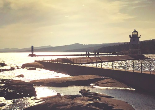 A serene waterfront scene featuring a lighthouse, pier, and distant mountains under a golden sky.