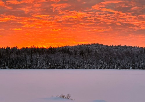 A vibrant sunset casts orange hues over a snowy landscape and forested hills, reflecting on a frozen lake.