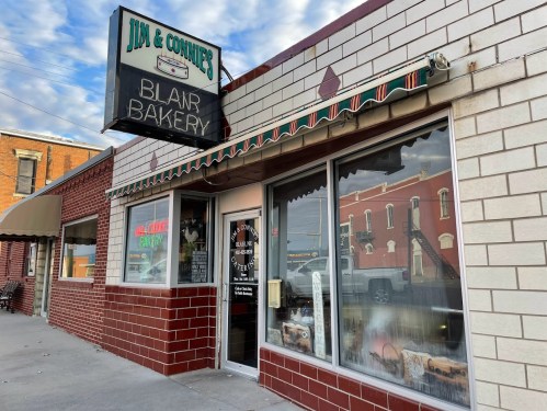 Exterior of Jim & Connie's Bakery, featuring a sign and large windows displaying baked goods, on a sunny day.