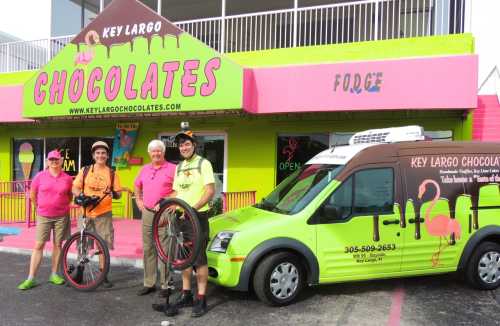 A group of four people in pink shirts stand outside a colorful chocolate shop with a green delivery van.