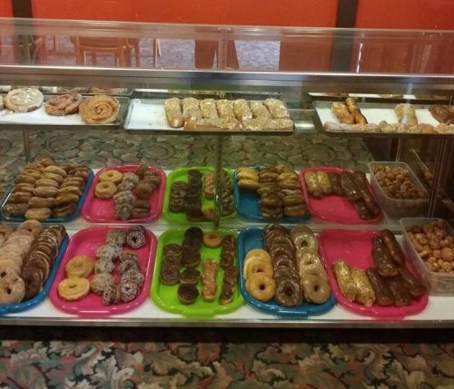 A display case filled with various types of donuts and pastries on colorful trays against a red wall.