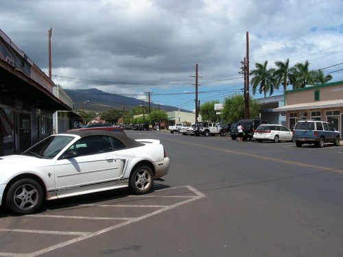 A quiet street scene with parked cars, shops, and palm trees under a partly cloudy sky. Mountains are visible in the background.