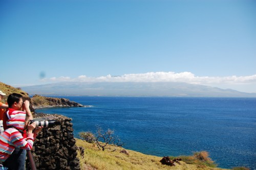 A child takes a photo of a scenic coastal view with blue water and distant mountains under a clear sky.